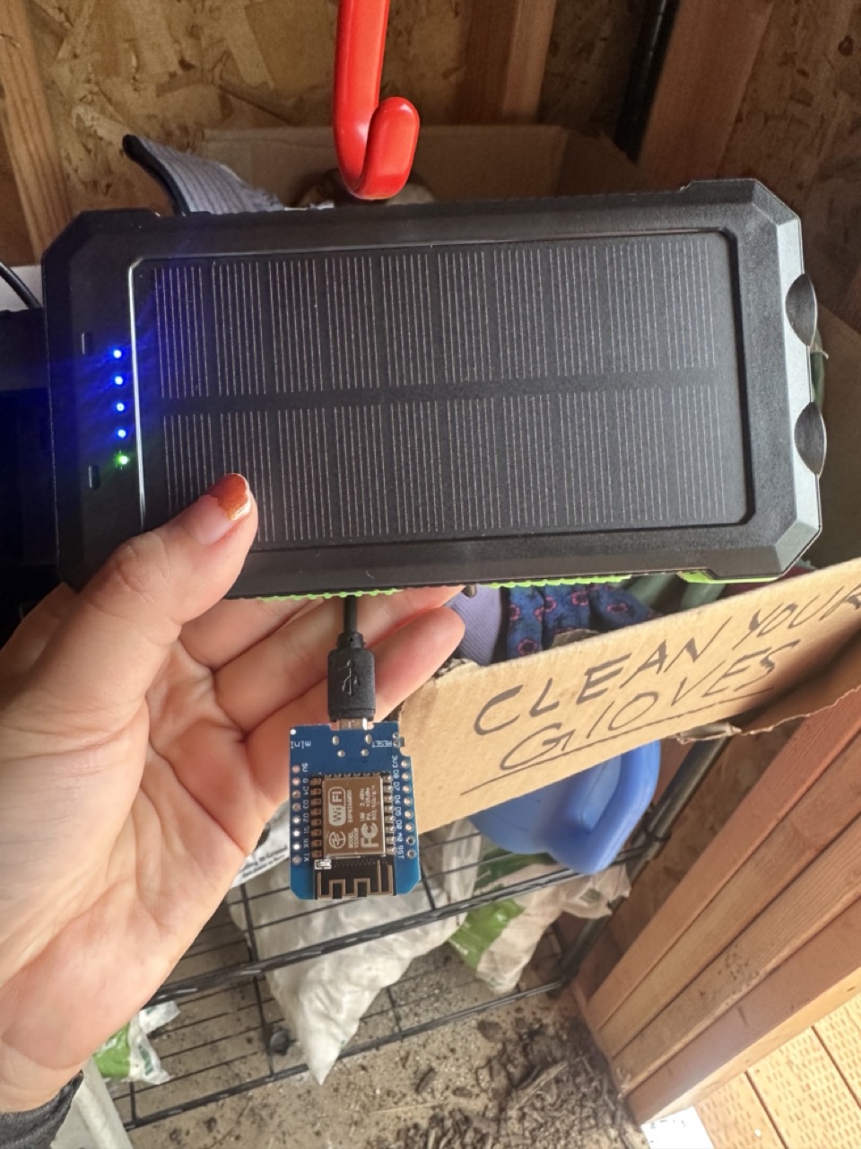 A hand is holding a charging electronic module connected to a portable solar panel with a sign in the background that reads "Clean your gloves."