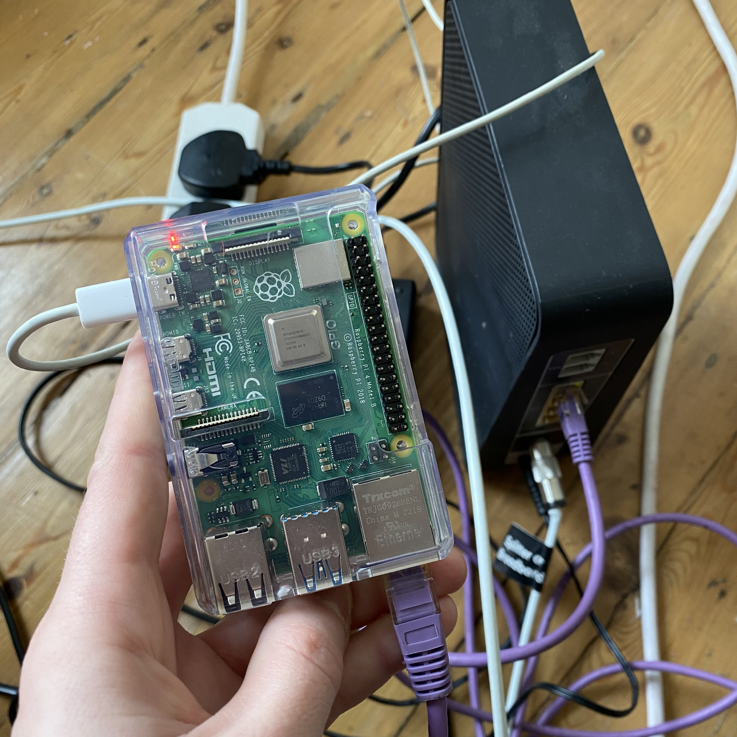A hand holds a transparent-cased Raspberry Pi connected by cables, with a router and other cables in the background on a wooden floor.