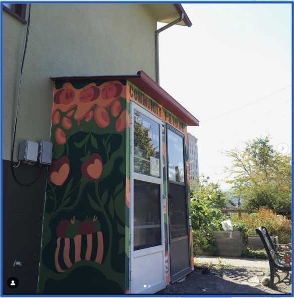 A small building with a mural of fruits and vegetables on its side, labeled "Community Fridge," stands next to a garden space and a bench.