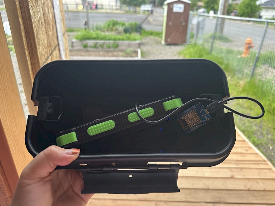 A person is holding an open black case containing an electronic device with green accents and a connected circuit board, with a garden and a porta-potty visible in the background.