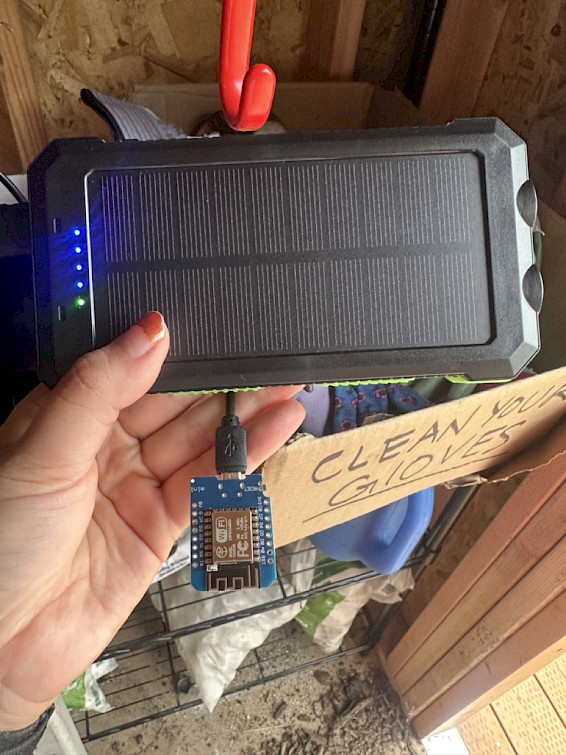 A hand is holding a charging electronic module connected to a portable solar panel with a sign in the background that reads "Clean your gloves."