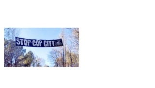 A banner reading "STOP COP CITY" hangs between trees under a clear sky.