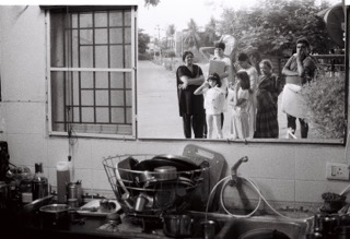 A black-and-white photo showing a cluttered kitchen sink with dirty dishes, viewed through an open window with a group of people standing outside and looking in.