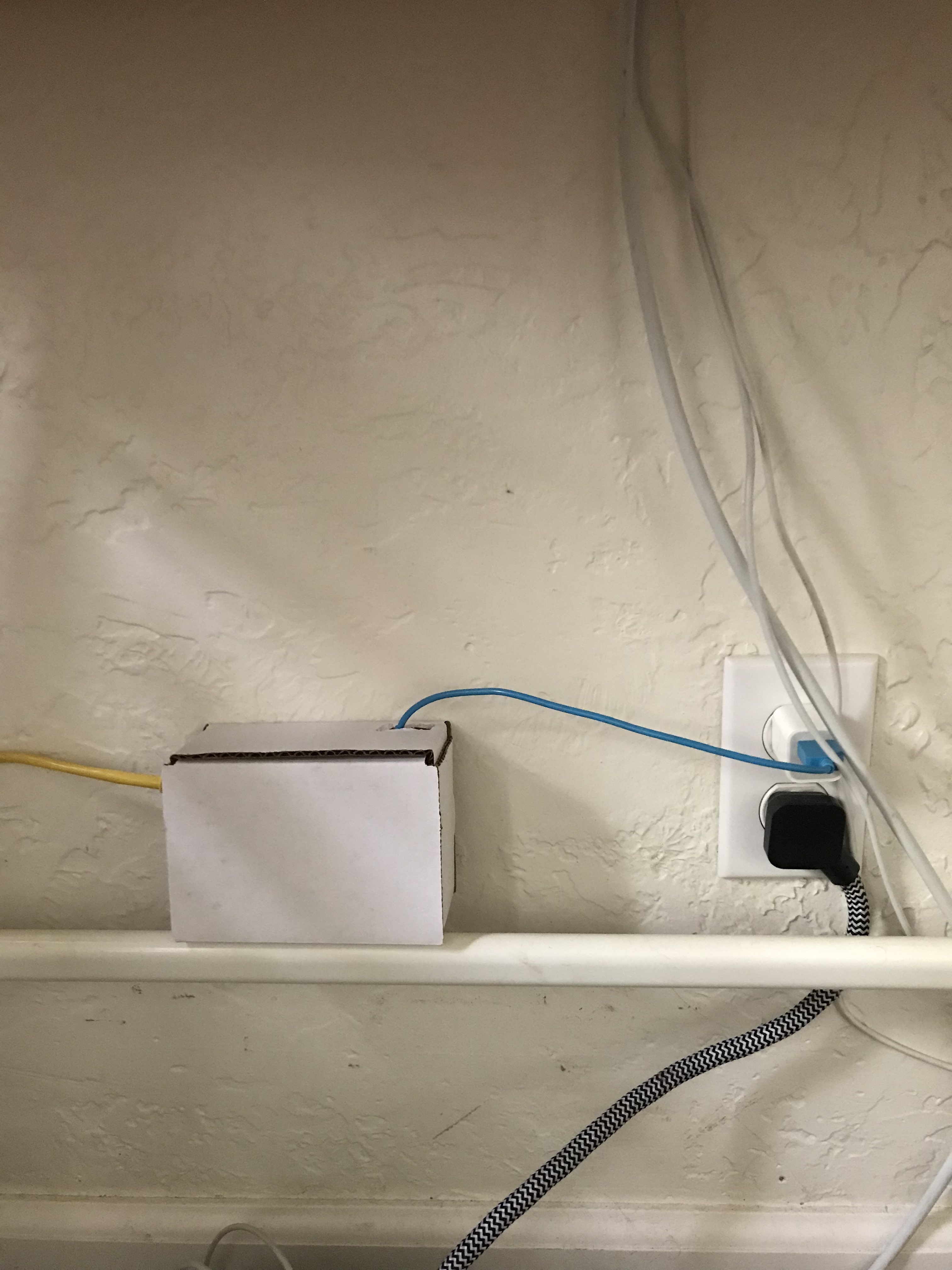 A white cardboard box with yellow and blue wires protruding from it sits on a shelf near an electrical outlet with two cords plugged in.