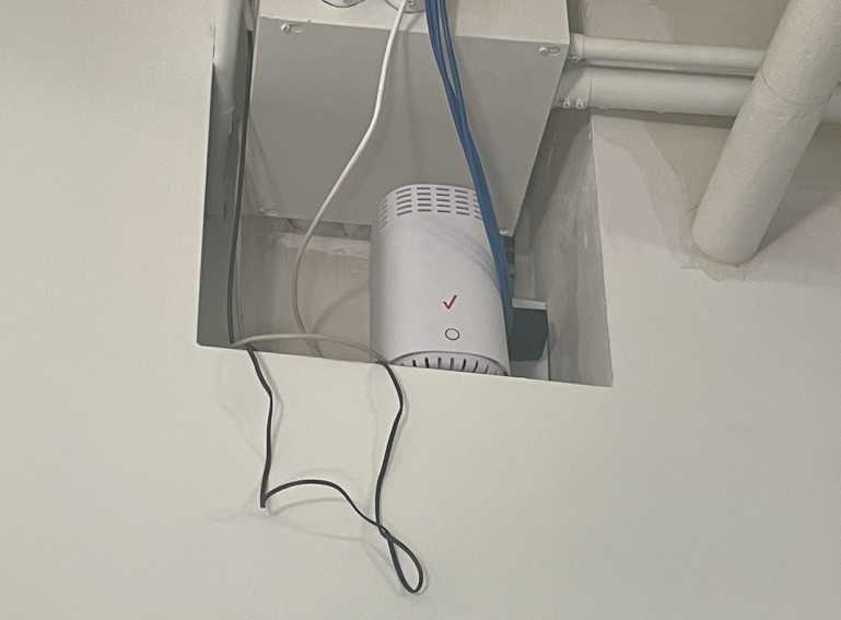 A white electronic device with a red check mark and cables is installed in a recessed section of a ceiling.