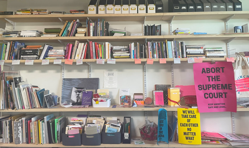 A wall of bookshelves filled with various books, folders, documents, and items with signs including "ABORT THE SUPREME COURT" and "WE WILL TAKE CARE OF EACH OTHER NO MATTER WHAT."