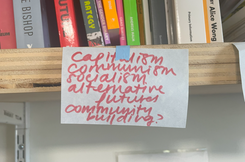 A bookshelf with various books and a sticky note with handwritten words: "Capitalism, Communism, Socialism, Alternative futures, Community building?"