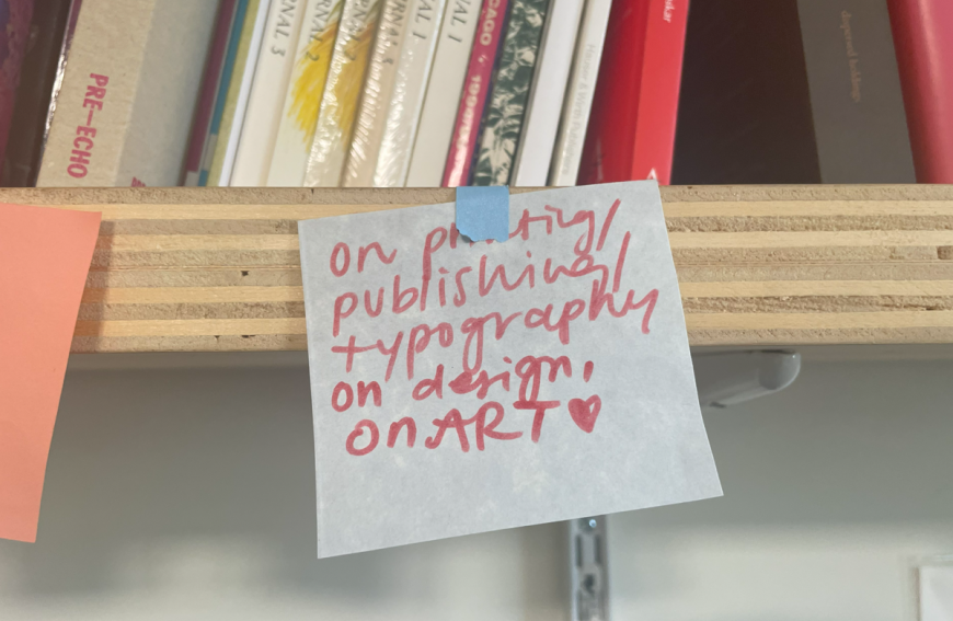 A handwritten note on blue tape reads "on print/publishing typography on design, on ART" with a heart symbol at the end, attached to a wooden shelf with books on the background.