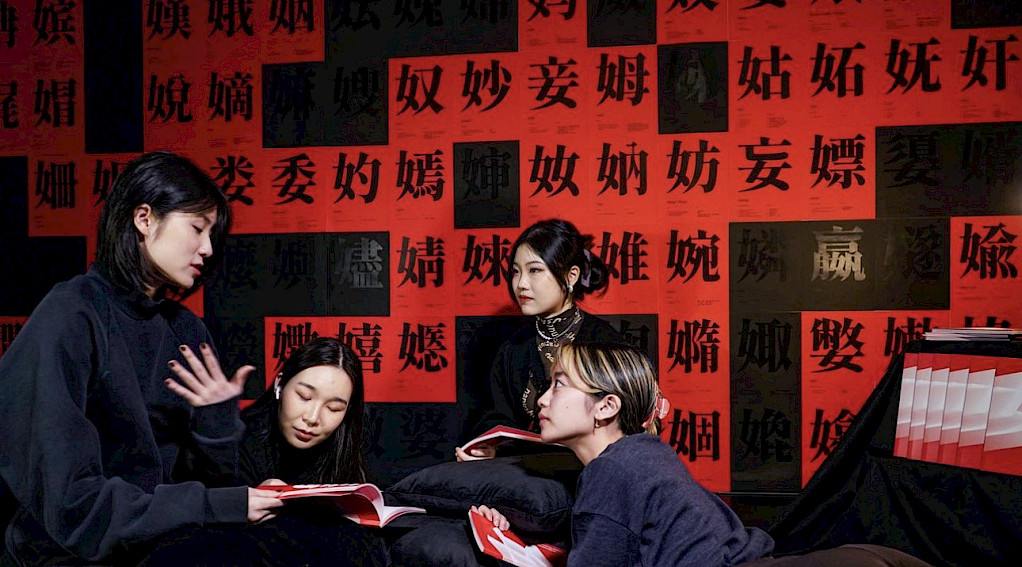 Four people are sitting and conversing in front of a wall covered in red and black posters with Chinese characters.