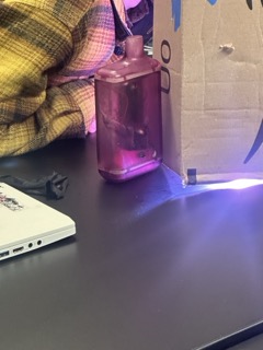 A person in a yellow plaid shirt is sitting at a desk with a purple bottle, a cardboard setup, and a laptop, with a bright light shining from underneath the cardboard.