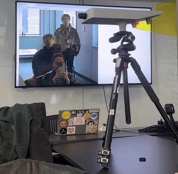 A TV screen displaying three people, one of whom is taking a selfie, is shown in an office setting with a tripod and a laptop featuring various stickers in the foreground.