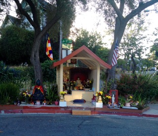 A small roadside shrine featuring a Buddha statue, surrounded by flowers, candles, and flags, is nestled between two trees.