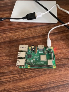 A Raspberry Pi single-board computer connected to a white USB cable is placed on a wooden surface, with an external hard drive in the background.