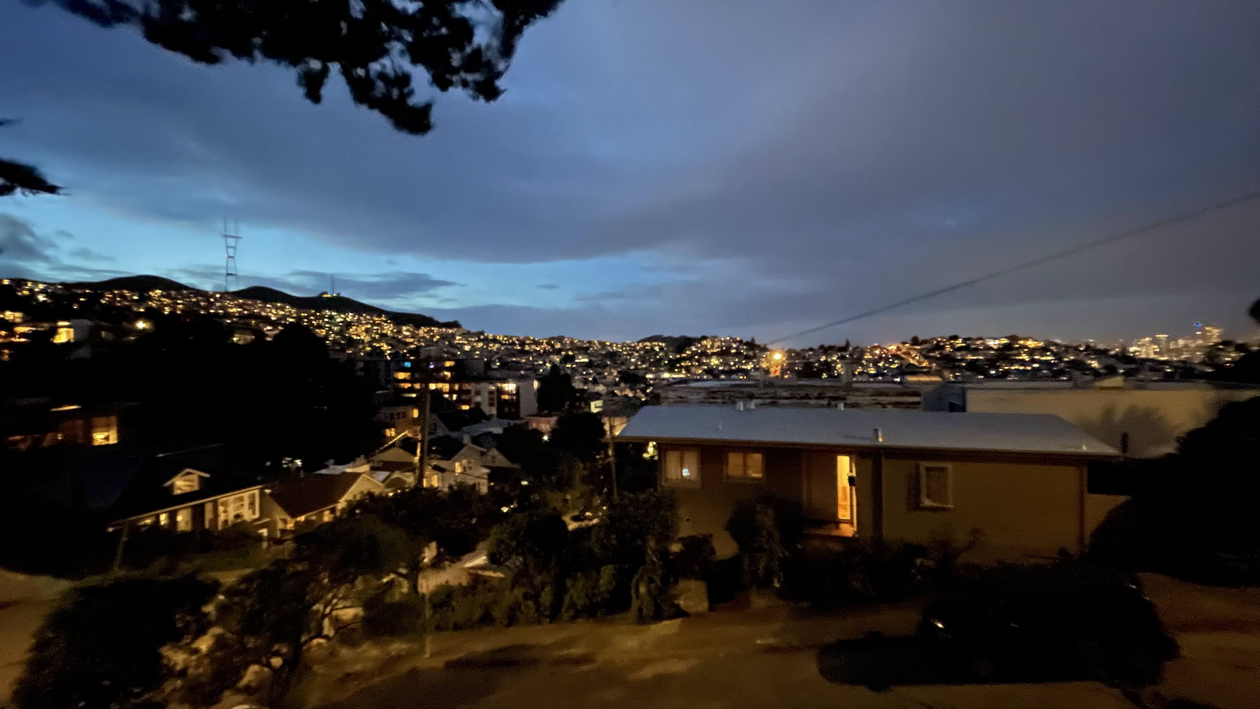 A cityscape at dusk shows a view of illuminated houses and buildings nestled among rolling hills, with a tower and distant lights under a partly cloudy sky.