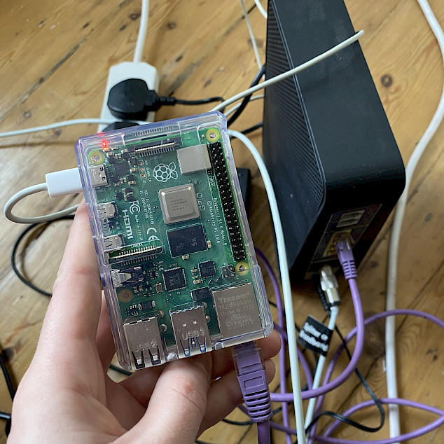 A hand holds a transparent-cased Raspberry Pi connected by cables, with a router and other cables in the background on a wooden floor.