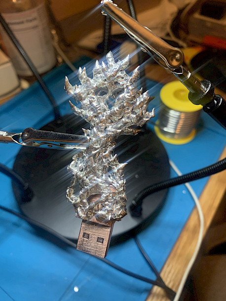 A metallic spiky object is secured by clips typically used for soldering, with a USB connector protruding from one end.