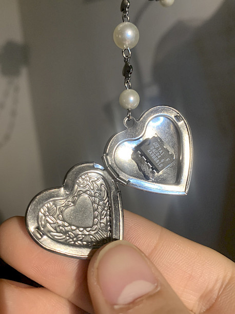 A close-up of a person's hand holding an open, heart-shaped locket with intricate designs and pearl decorations on the chain.